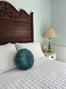 A wooden headboard behind the white linens and blue round pillow. A small lamp sits on a white dresser in the corner.