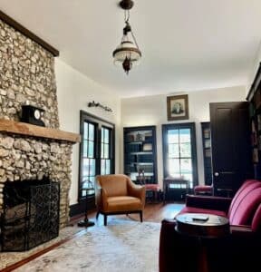 A rock fireplace on the left with windows and bookcases near the center. A red velveteen couch with a small table are on the right of the image.