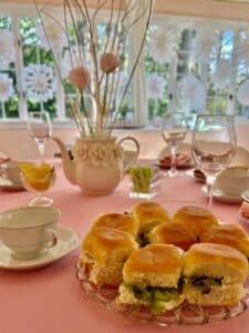 Sandwiches in the foreground on a pink tablecloth with teacups, glasses and a teapot with pink flowers and silver sticks. 