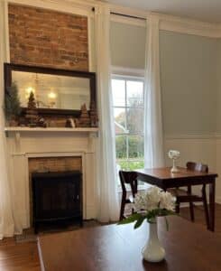 Dining room with wood tables and white flower arrangements. Fireplace with mirror and Christmas decorated mantle. 