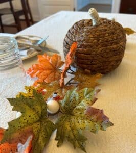 Brown wicker pumpkin in front of orange and green leaves on set table. 