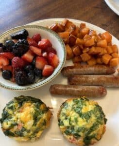 Plate with bowl of fruit, side of orange sweet potatoes, three sausage links and two round egg puffs. 