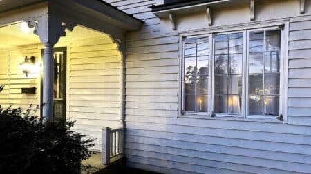 White house with porch in the evening. Porch light on left of image and windows on right side.