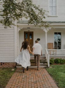 Lady in white dress with man in white shirt holding hands and walking up porch on white house.