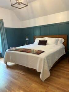 Bed with white blanket and quilt. Wooden floor with blue wainscoting wall and white on top. 