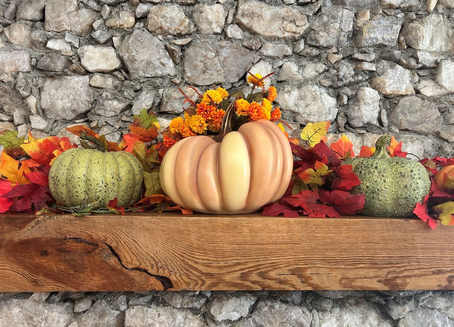 Green and orange pumpkins on wooden mantle. Rock background and some fall leaves of orange, red, and yellow.
