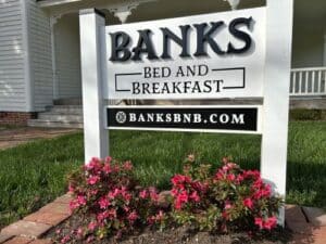 White with black lettering Banks Bed and Breakfast sign. Pink azalea bushes under sign.