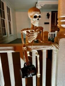 Skeleton holding black mug as it leans over a white railing. 