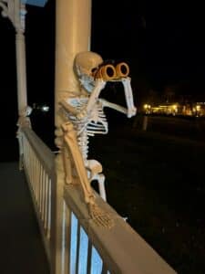 Skeleton sitting on white railing looking through orange binoculars.