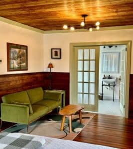 Green couch on wooden floor with glass doors in background. Pictures on the wall and small wooden table. 