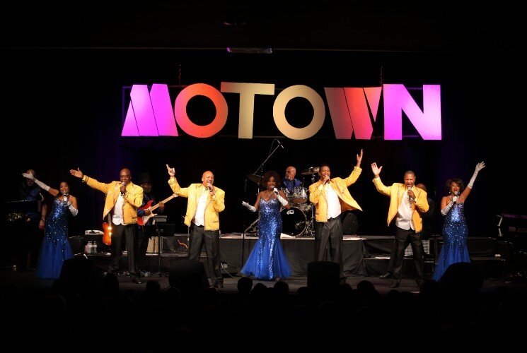Three women in blue dresses and four men in yellow suit coats singing on stage with a Motown sign