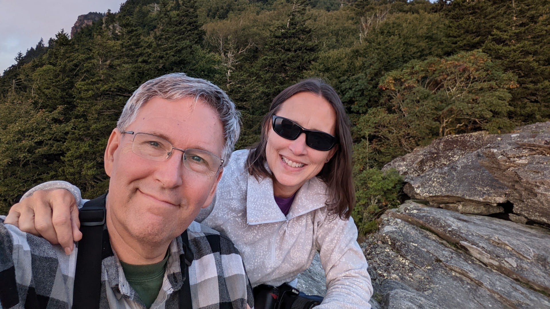 A man in a plaid shirt with a woman in sunglasses sitting atop a cliff's edge surrounded by trees
