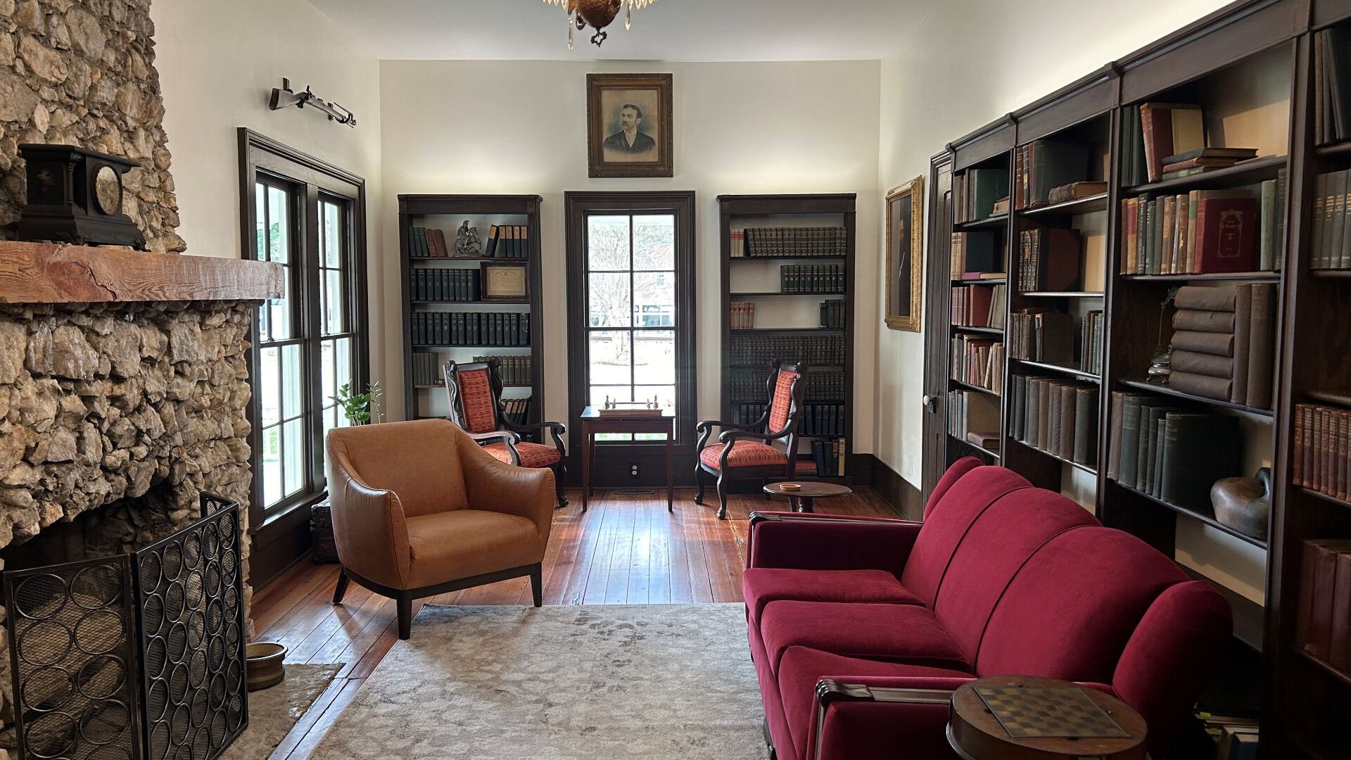Red couch in front of a bookcase filled with books. More bookcases on each side of a window and another tan chair. The library at Banks Bed and Breakfast.