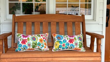 A wood bench with two colorful floral pillows outside on a patio by a wooden door and window