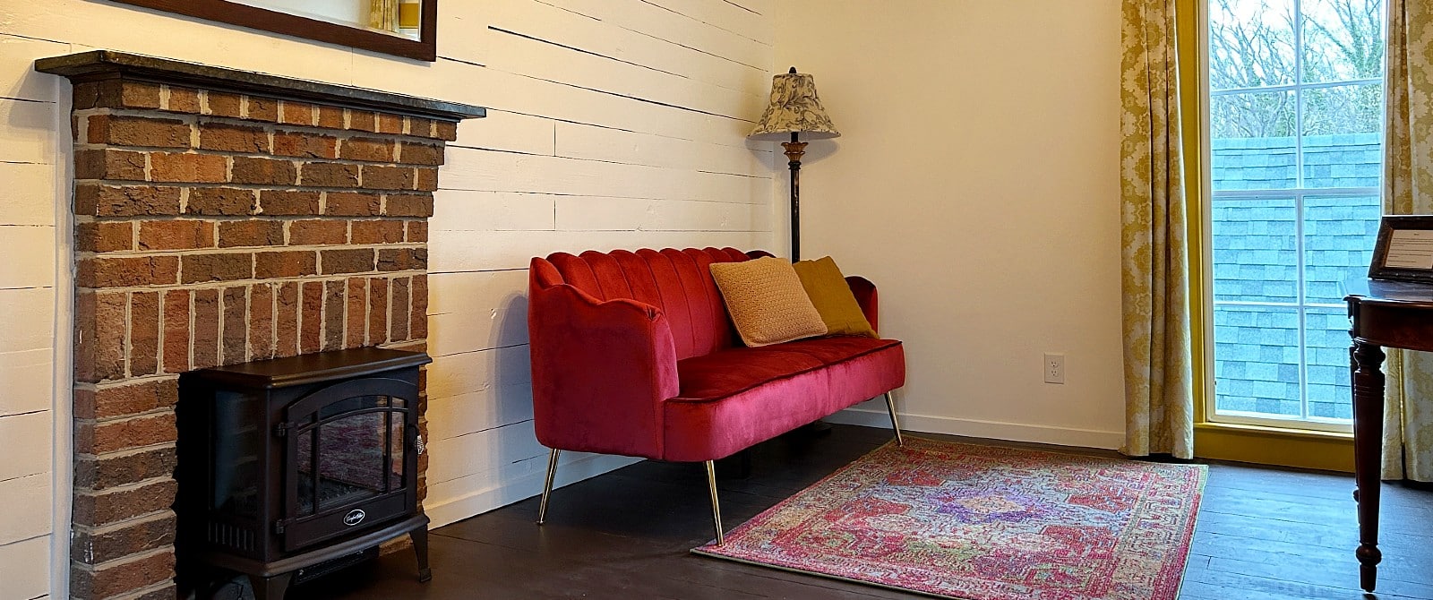 Gorgeous red velvet couch with a rug by a brick fireplace and large window with floral curtains