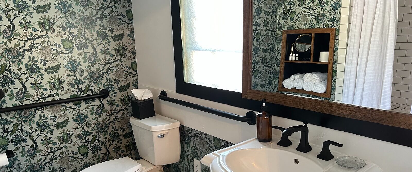 Bathroom with pedestal sink, large wood-framed mirror, toilet below a window and floral wallpaper
