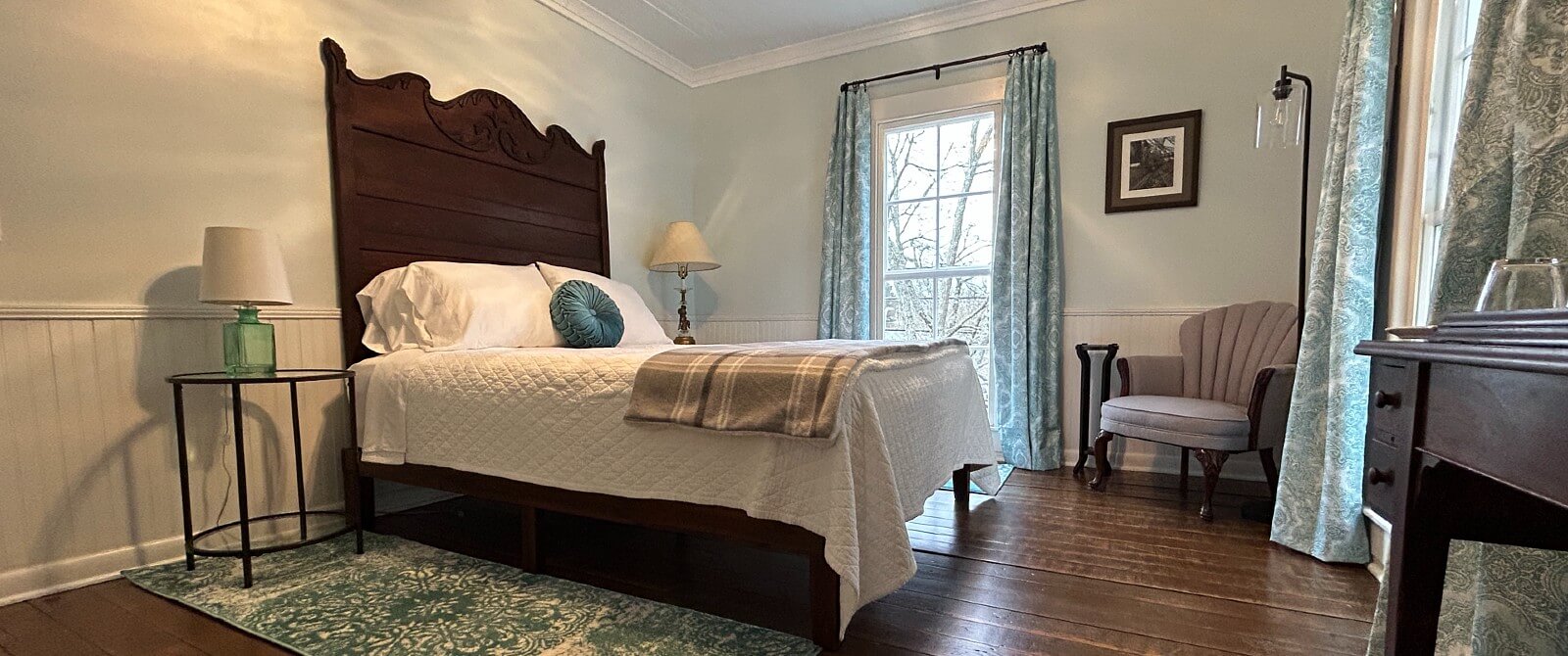 Guest room with queen bed with large antique headboard, side tables with lamps and large windows with floral curtains