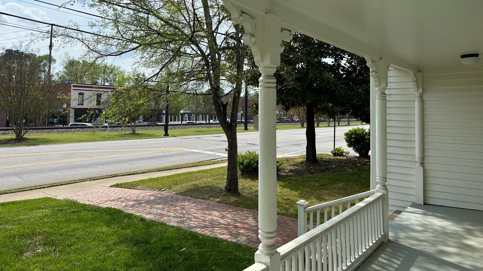 A front porch of a white home facing a street with a row of buildings beyond some trees