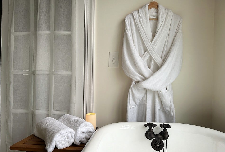 A plush white robe hanging by a claw foot tub with two rolled towels on a wooden side table