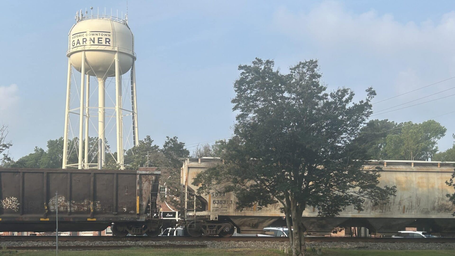 Water tower with brown and cream trainncars on the railroad track and one tree.